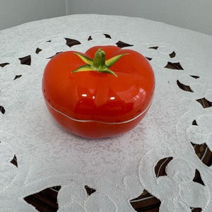 May include: A red ceramic tomato shaped dish with a green stem and leaves. The dish is sitting on a white lace tablecloth.