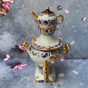 May include: A white porcelain samovar with gold and blue accents. The samovar has a round body with a spout on one side and a handle on the other. The top of the samovar is decorated with a smaller teapot with a gold lid and handle.