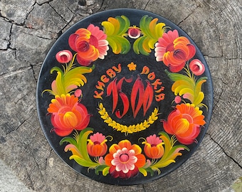 May include: A black wooden plate with a floral design painted in red, orange, yellow, and green. The plate has the text "Kiev" and "Dokib" written in a circle around the center.