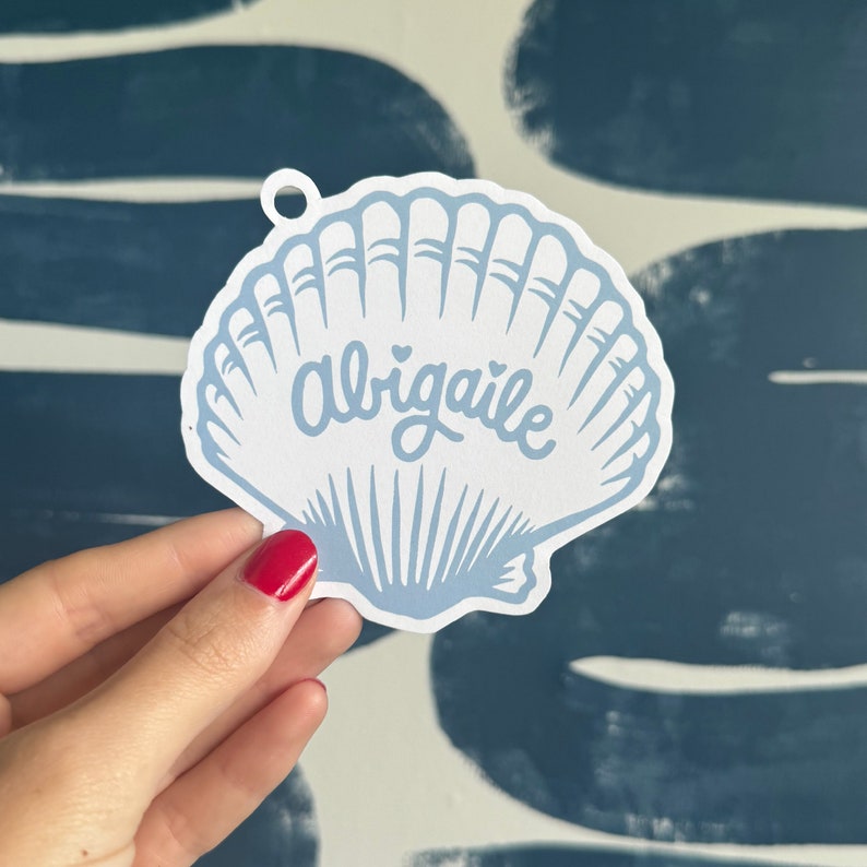 Puede incluir: Una pegatina en forma de concha marina azul claro y blanca con el nombre "Abigale" en escritura cursiva. La pegatina tiene un peque&ntilde;o agujero en la parte superior para colgarla. El dise&ntilde;o presenta un borde festoneado y est&aacute; sujeta por una mano con esmalte de u&ntilde;as rojo.