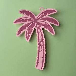 May include: A pink paper palm tree decoration with the name "Taylor" written on the trunk. The palm tree is cut out with a loop at the top for hanging. The background is a solid light green.