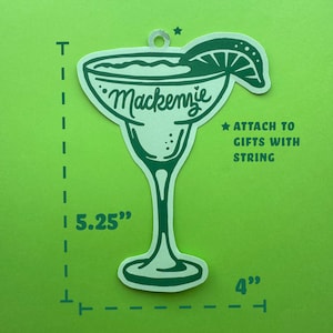 May include: A green margarita-shaped gift tag with the name "Mackenzie" in cursive. The tag is 5.25 inches tall and 4 inches wide, with a lime wedge and a hole for string. Text on the tag reads "Attach to gifts with string."