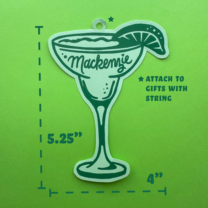 May include: A green margarita-shaped gift tag with the name "Mackenzie" in cursive. The tag is 5.25 inches tall and 4 inches wide, with a lime wedge and a hole for string. Text on the tag reads "Attach to gifts with string."