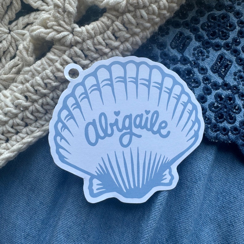 Puede incluir: Una pegatina en forma de concha de color azul claro con el nombre "Abigale" en escritura cursiva. La pegatina tiene un peque&ntilde;o agujero en la parte superior para colgarla. La pegatina est&aacute; sobre una superficie de mezclilla azul, con un art&iacute;culo de ganchillo color crema y una tela bordada azul en el fondo.
