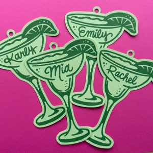 May include: Green margarita-shaped paper cutouts with names: Karly, Emily, Mia, and Rachel. Each cutout has a lime wedge detail and a small hole at the top. The cutouts are arranged on a bright pink background.