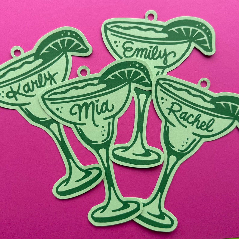 May include: Green margarita-shaped paper cutouts with names: Karly, Emily, Mia, and Rachel. Each cutout has a lime wedge detail and a small hole at the top. The cutouts are arranged on a bright pink background.