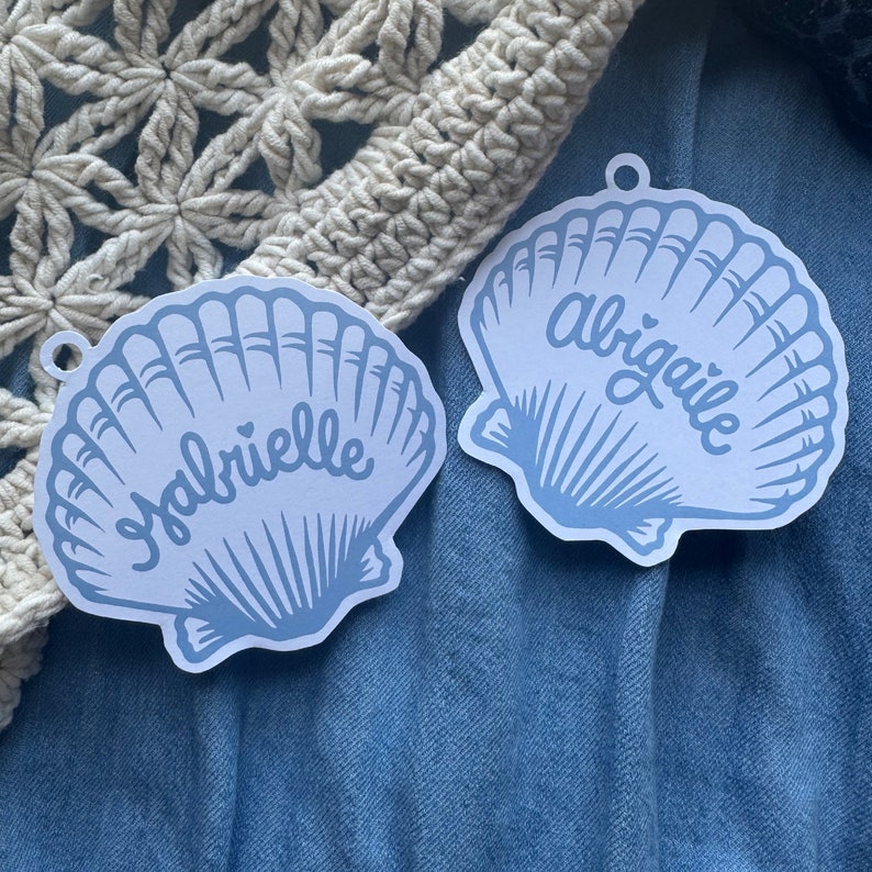 Puede incluir: Dos adornos en forma de concha de color azul claro con los nombres "Gabrielle" y "Abigale" en escritura cursiva. Cada adorno tiene un peque&ntilde;o agujero en la parte superior para colgar. El fondo incluye un art&iacute;culo de ganchillo color crema y tela vaquera azul.