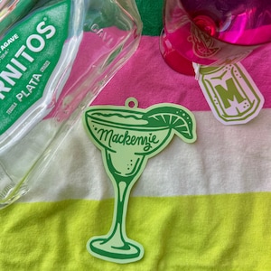 May include: A light green margarita-shaped ornament with the name "Mackenzie" in cursive. The ornament has a lime wedge and is on a striped pink and yellow towel. A bottle of tequila and a pink drink are also visible.