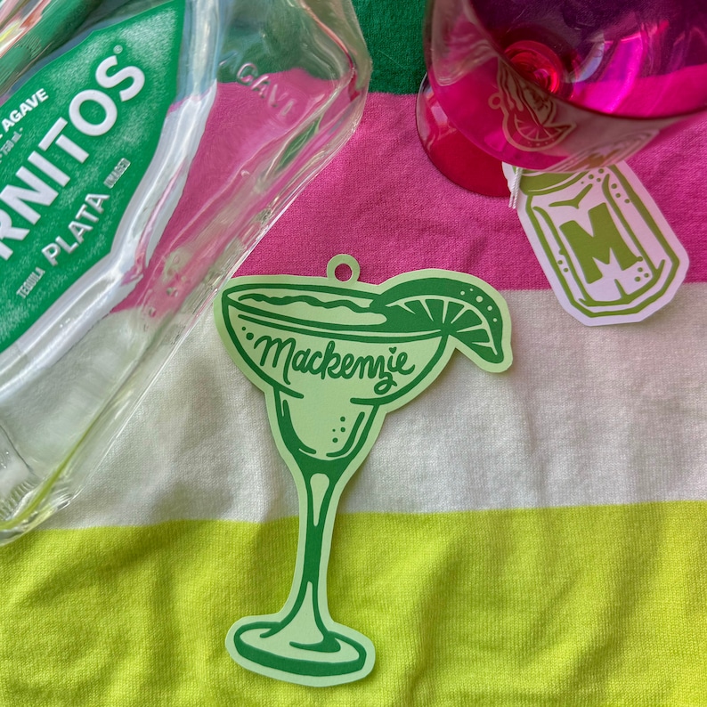 May include: A light green margarita-shaped ornament with the name "Mackenzie" in cursive. The ornament has a lime wedge and is on a striped pink and yellow towel. A bottle of tequila and a pink drink are also visible.