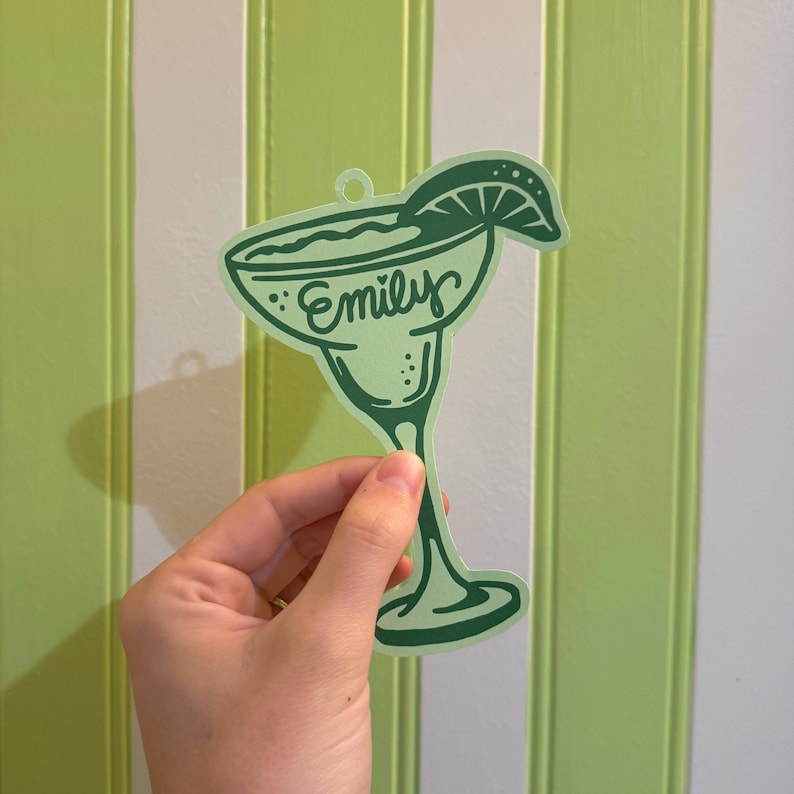 May include: A light green margarita glass-shaped ornament with the name "Emily" in script. The ornament has a lime wedge and is held against a green and white striped background.