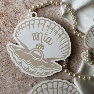 May include: Off-white seashell-shaped tags with the name "Mia" in cursive, a ring graphic, and a pearl necklace. The tags have a beige outline and are on a textured surface.