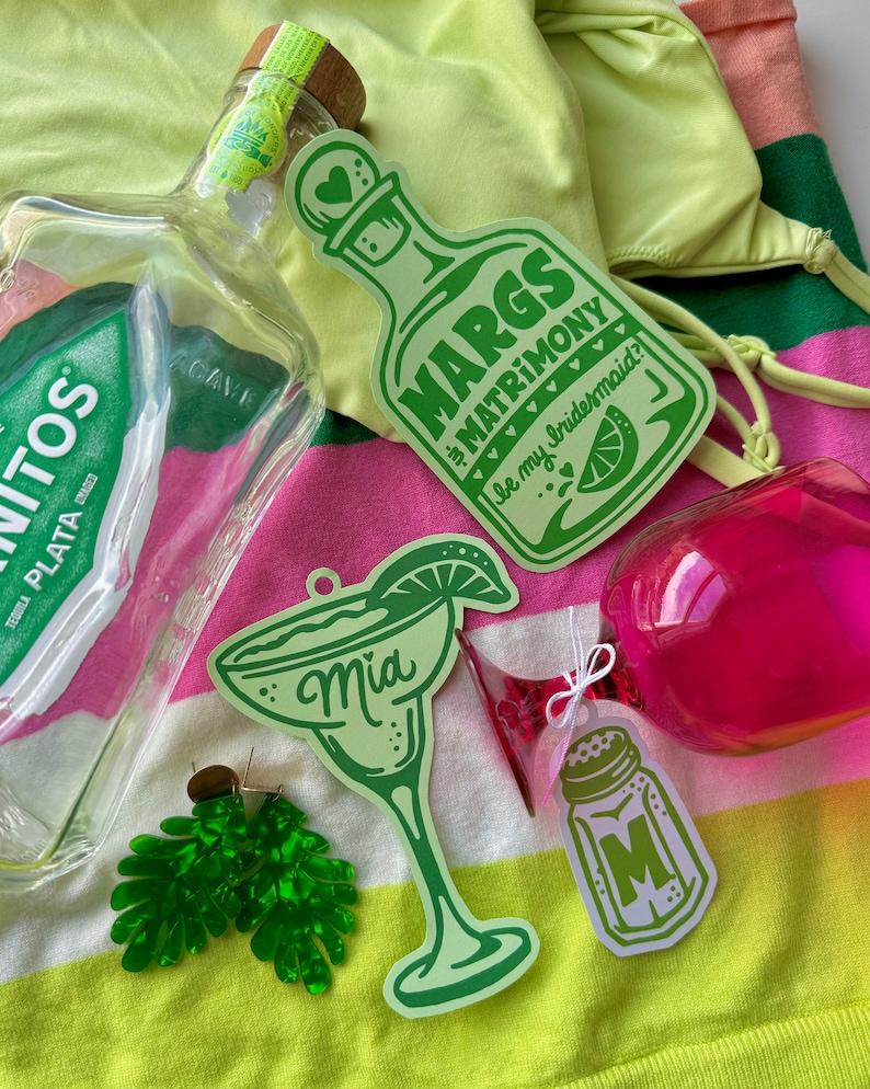 May include: A collection of margarita-themed items, including a tequila bottle, green margarita-shaped stickers with text, a pair of green leaf earrings, and a pink purse, set against a striped background.