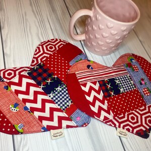 May include: Two red heart-shaped pot holders with a patchwork design of red, white, and blue fabric. The pot holders are made of different patterns, including polka dots, stripes, and geometric shapes. The pot holders are on a white wooden surface.