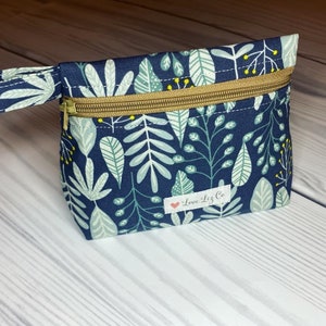 May include: A small blue zippered pouch with a white and green leafy pattern. The pouch has a gold zipper and a handle. The pouch has a small red heart with the text "Love Liz Co." on it.
