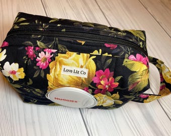 May include: A black quilted floral print wipe case with a zipper closure and a handle. The case features yellow and pink flowers with the text "Love Liz Co" on a white label. A Huggies wipe dispenser is visible.