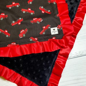 Pu&ograve; includere: Una coperta per bambini con un tessuto nero a fantasia di camioncini rossi. Ha un bordo a volant in raso rosso e un tessuto nero strutturato. L'etichetta recita "Love Liz co."