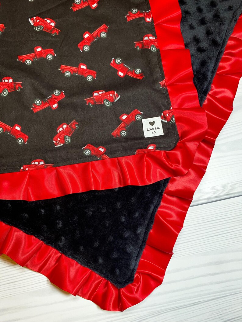 Pu&ograve; includere: Una coperta per bambini con un tessuto nero a fantasia di camioncini rossi. Ha un bordo a volant in raso rosso e un tessuto nero strutturato. L'etichetta recita "Love Liz co."