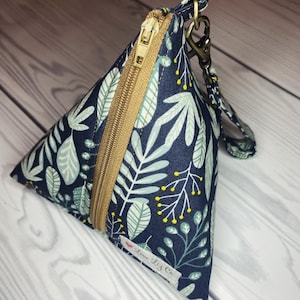May include: A small, triangular zippered pouch with a blue background and a white and green floral pattern. The pouch has a gold zipper and a gold chain attached to a brown strap. The pouch has a small label that says "Love Liz Co."