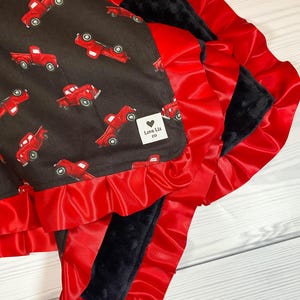 Pu&ograve; includere: Una coperta per bambini con un motivo di camion rossi su sfondo marrone scuro. La coperta &egrave; rifinita con un volant di raso rosso e ha un retro in peluche nero. L'etichetta recita "Love Liz co."