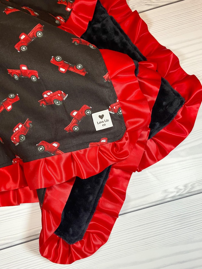 Pu&ograve; includere: Una coperta per bambini con un motivo di camion rossi su sfondo marrone scuro. La coperta &egrave; rifinita con un volant di raso rosso e ha un retro in peluche nero. L'etichetta recita "Love Liz co."