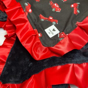 Pu&ograve; includere: Una coperta per bambini con sfondo nero e motivo di camion rossi. La coperta ha un bordo in raso rosso con bordo a balze e un retro in peluche nero. &Egrave; visibile una piccola etichetta bianca con la scritta "Love Liz co".