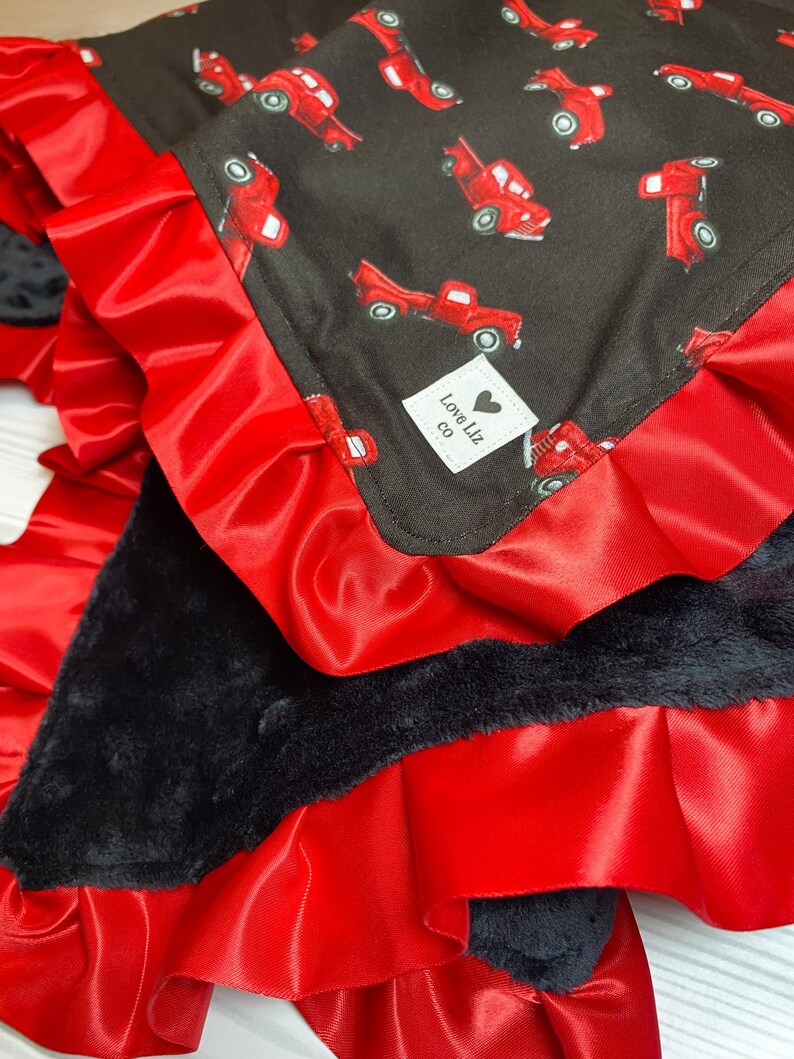 Pu&ograve; includere: Una coperta per bambini con sfondo nero e motivo di camion rossi. La coperta ha un bordo in raso rosso con bordo a balze e un retro in peluche nero. &Egrave; visibile una piccola etichetta bianca con la scritta "Love Liz co".