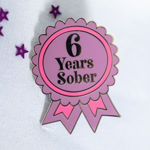 Könnte beinhalten: Ein lila-rosa Emaille-Anstecknadel mit dem Text "6 Years Sober". Die Anstecknadel hat die Form eines Bandes mit einem gewellten Rand und zwei rosa Bändern am unteren Rand. Lila sternförmige Konfetti sind um die Anstecknadel verstreut.