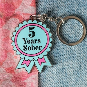 May include: A silver keychain with a blue and pink enamel ribbon featuring the text "5 Years Sober".