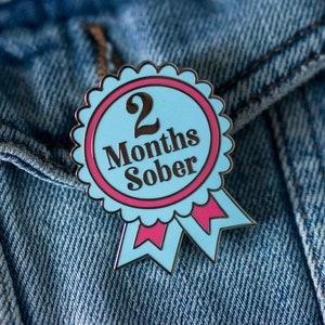 May include: A blue and pink enamel pin with a white ribbon. The pin says "2 Months Sober".