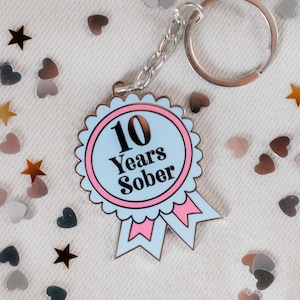 May include: A blue and pink enamel keychain with a white ribbon and the text "10 Years Sober".