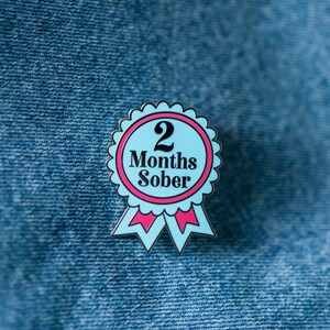 May include: A blue enamel pin with a pink ribbon and the text "2 Months Sober".