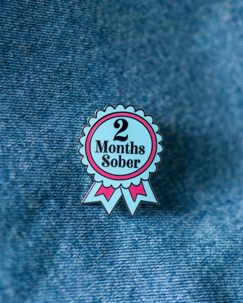 May include: A blue enamel pin with a pink ribbon and the text "2 Months Sober".