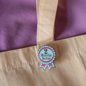 May include: A blue and pink enamel pin with a white ribbon and the text "2 Months Sober".
