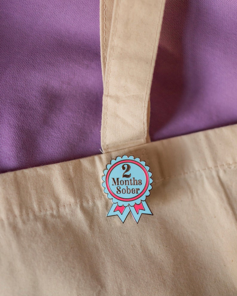 May include: A blue and pink enamel pin with a white ribbon and the text "2 Months Sober".