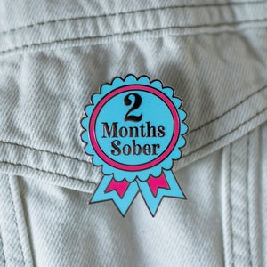 May include: A blue and pink enamel pin with a white ribbon. The pin features a white circle with the text "2 Months Sober" in black.