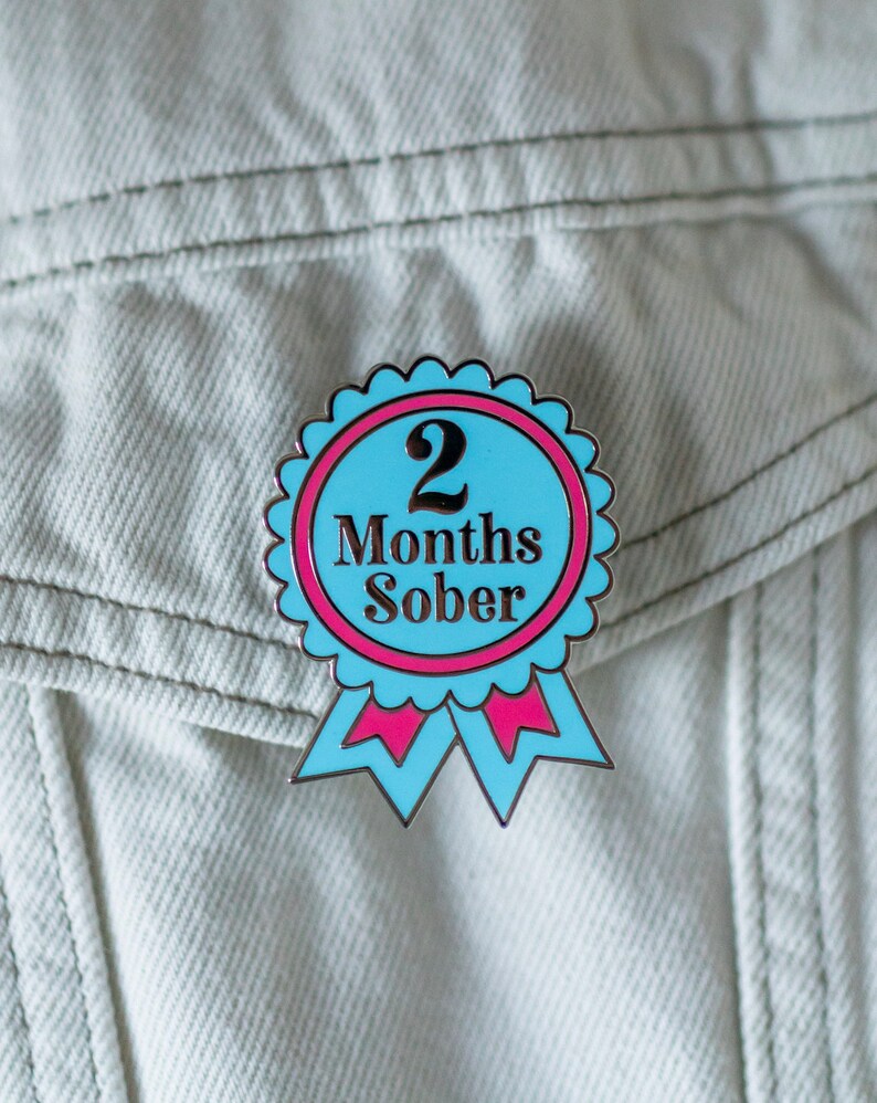 May include: A blue and pink enamel pin with a white ribbon. The pin features a white circle with the text "2 Months Sober" in black.