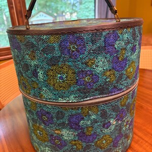 Pode incluir: Uma mala de toucador cilíndrica com um exterior de tecido texturizado com um padrão floral repetitivo em tons de azul, verde-azulado e verde-azeitona. A mala inclui uma pega superior castanha escura, um fecho de correr e rebordos escuros.