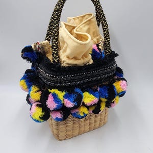 May include: A woven straw tote bag with a black handle and a colorful pom-pom trim. The bag has a black and white trim with sequins and a beige fabric lining.