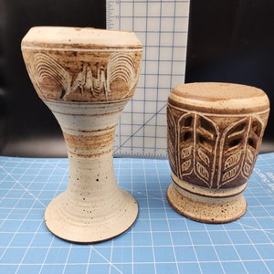 Puede incluir: Dos candelabros de cerámica con esmalte marrón y blanco. El candelero más alto tiene una base ancha y una parte superior acampanada con un diseño tallado. El candelero más bajo tiene una forma cilíndrica con un diseño tallado de hojas.