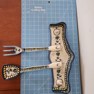 May include: A ceramic spoon and fork set with a decorative holder. The utensils have a blue, yellow, and white floral design. The holder is cream-colored with a blue border and grape vine pattern. The set is on a blue cutting mat.
