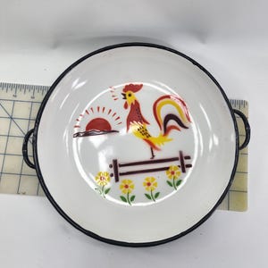 Puede incluir: Plato de esmalte blanco con borde y asas negros. Presenta una colorida ilustración de un gallo, un sol naciente, una valla de madera y flores amarillas. Un artículo de cocina vintage, de unos 20 cm de diámetro.