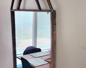 May include: A wooden framed arched mirror with a geometric design. The mirror reflects a room with a wooden table and green chairs.