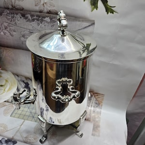 May include: A polished silver-tone metal beverage dispenser with a cylindrical body, a removable lid featuring a decorative finial, ornate side handles, a small spigot, and four claw-style feet. The surface is highly reflective.