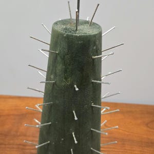 May include: A dark green, cylindrical sculpture with numerous silver nails protruding from its surface. The sculpture has a slightly conical shape and stands on a wooden surface. A single, taller nail is centered on top.
