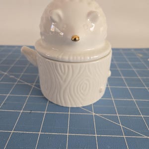 May include: White ceramic hedgehog shaped jar with a lid. The lid is shaped like a tree stump with a wood grain pattern. The hedgehog has a gold nose.