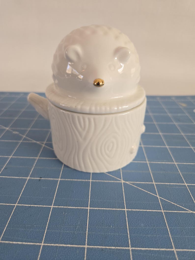 May include: White ceramic hedgehog shaped jar with a lid. The lid is shaped like a tree stump with a wood grain pattern. The hedgehog has a gold nose.