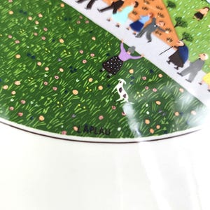May include: A vintage ceramic plate featuring a green grassy field, a white path, and a group of people walking. The plate has a white border with the word "LAPLAU" in black lettering.