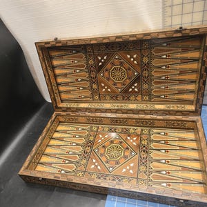 Op de afbeelding: Open houten backgammonbord met ingelegde geometrische patronen in bruin, beige en crème. Het rechthoekige bord heeft een scharnierend deksel, met gedetailleerd vakmanschap en een klassiek spelontwerp.