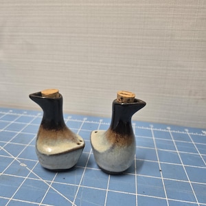May include: Two ceramic duck-shaped salt and pepper shakers. The shakers have a black head and neck, transitioning to brown and then a light gray body. Each shaker has a cork stopper.
