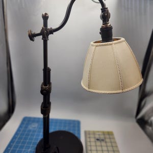 May include: A black metal desk lamp with a curved arm and a white fabric lampshade. The lamp has a round base and a decorative design on the arm.